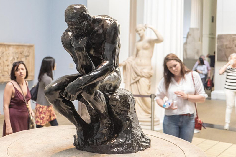 The image features a bronze sculpture of a deep-thinking figure, surrounded by museum visitors, emphasizing reflection and contemplation.