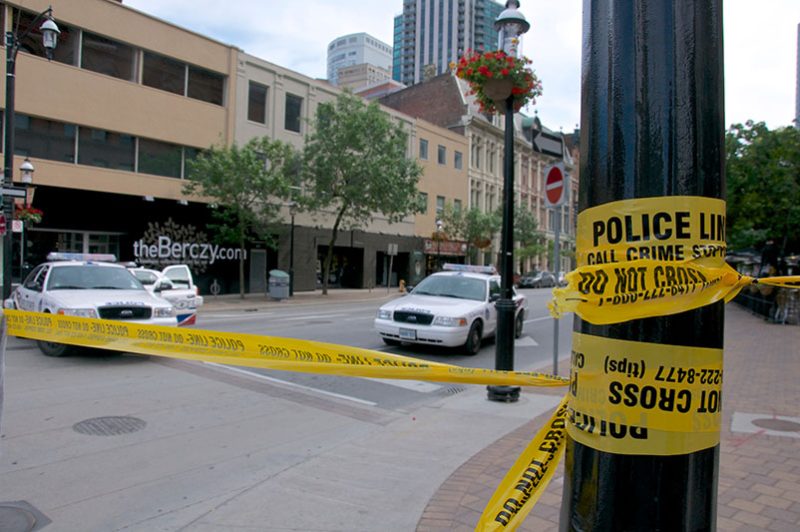 A street scene with police cars and yellow "POLICE LINE DO NOT CROSS" tape barricading an area. It conveys a sense of ongoing investigation or crime scene presence.